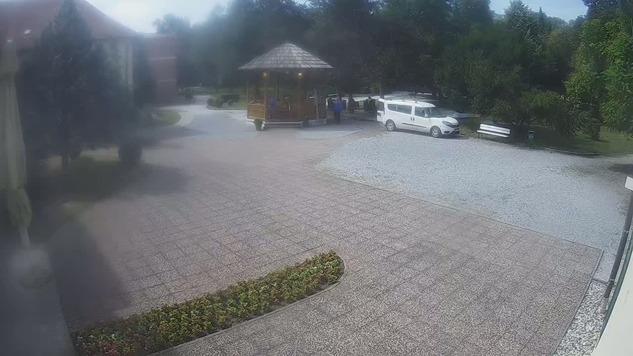 A paved courtyard with a wooden gazebo, a white van, and a park-like setting under an overcast sky.