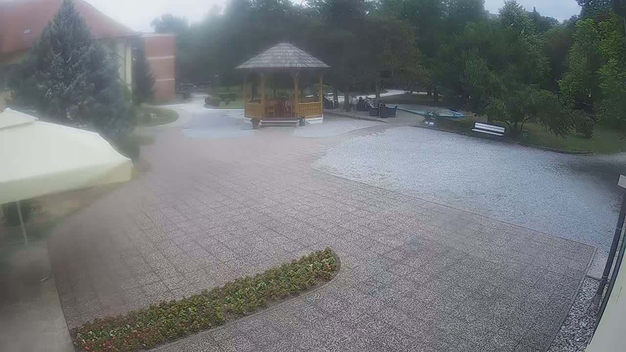 A paved courtyard with a wooden gazebo, a white van, and a park-like setting under an overcast sky.