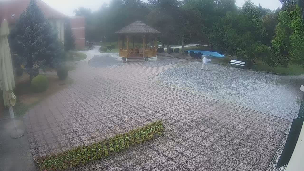 A person walks across a gravel pathway toward a gazebo in a courtyard, surrounded by brick paving, lush greenery, and buildings, on an overcast day.