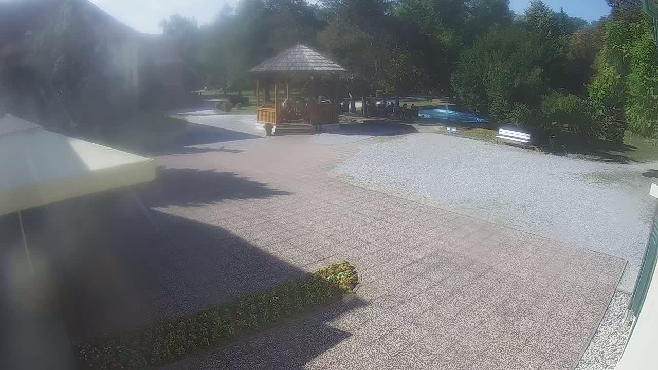 A paved outdoor area with a wooden gazebo, a gravel section, a bench, some landscaping, and a portion of a patio umbrella is visible on a sunny day.