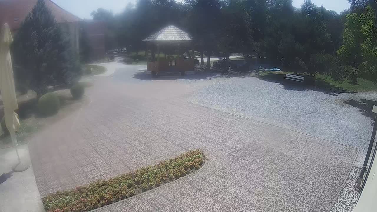 A wooden gazebo sits in a gravel courtyard on a sunny day, surrounded by green trees and landscaping.