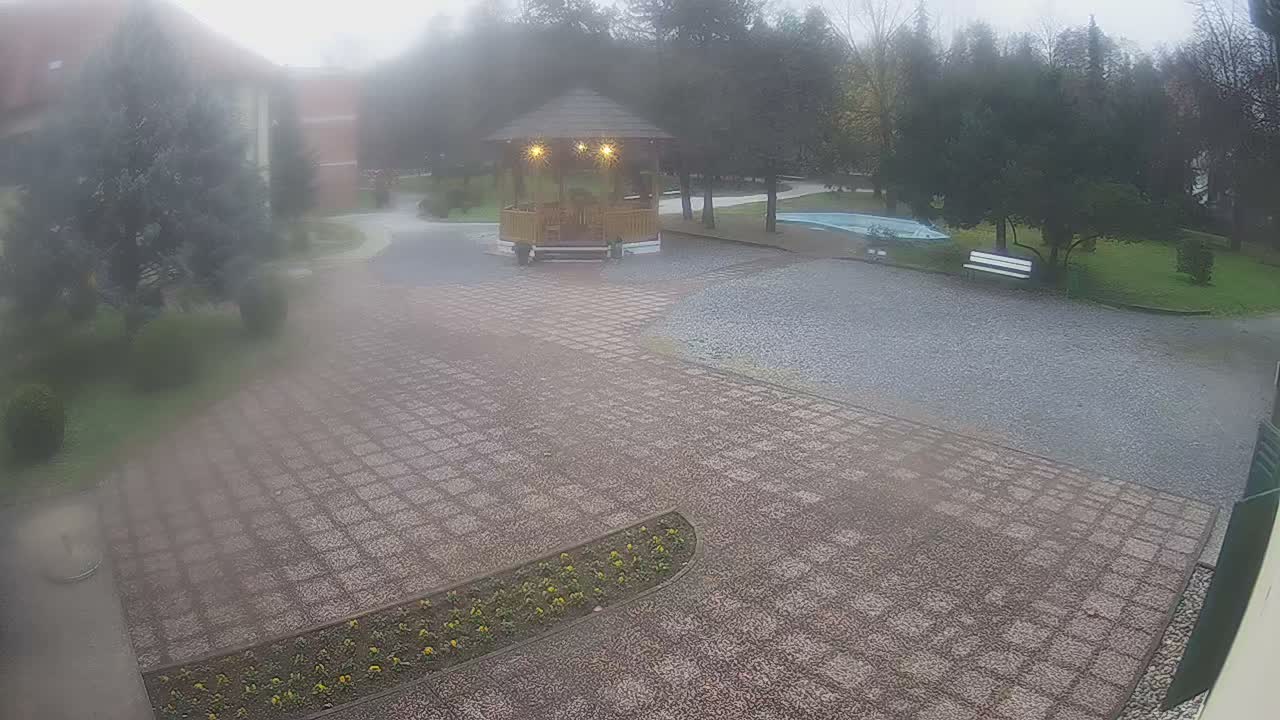A wooden gazebo sits in a gravel courtyard on a sunny day, surrounded by green trees and landscaping.