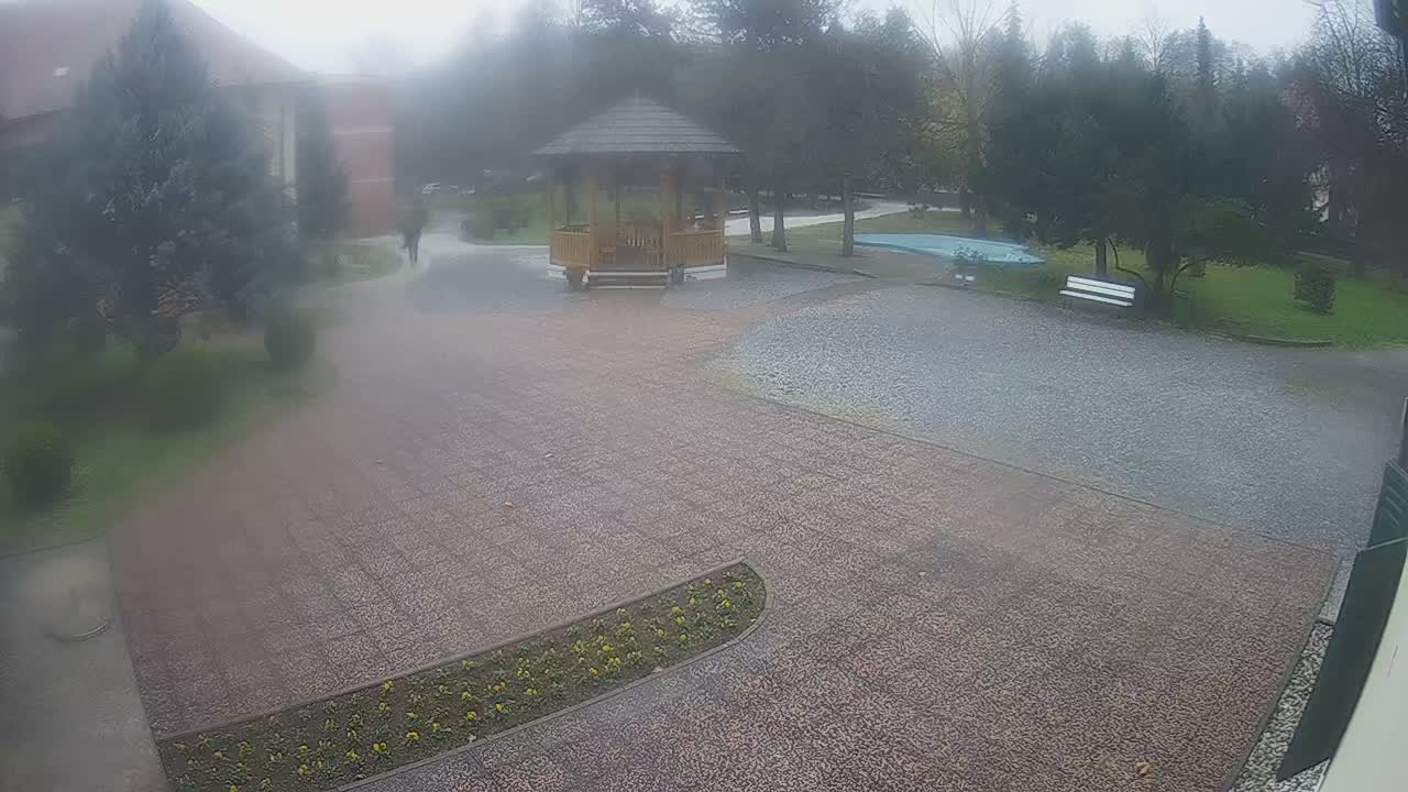 A misty, overcast outdoor scene reveals a paved courtyard with a central wooden gazebo, surrounded by lush trees, a small blue pool, a white bench, and a blurry figure walking towards a distant building.