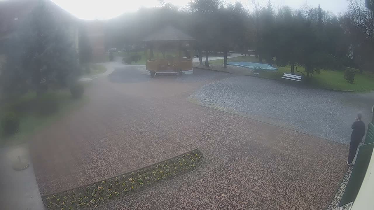 An elevated view shows an outdoor resort-like landscape with a paved courtyard, a central wooden gazebo, a distant swimming pool, and a person on the right talking on a phone, all under an overcast and hazy sky.