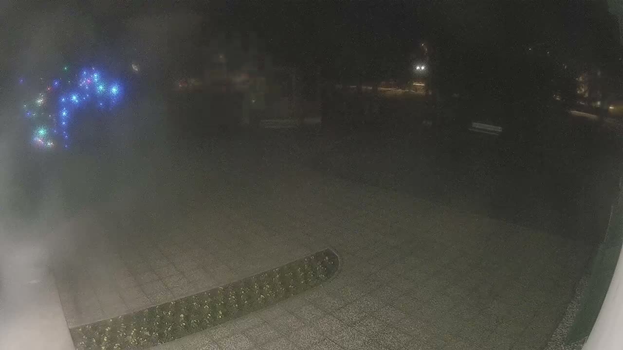 A dark, foggy night scene reveals a paved outdoor area with a planter, a blurry display of colorful lights on the left, and distant streetlights illuminating a dark park.