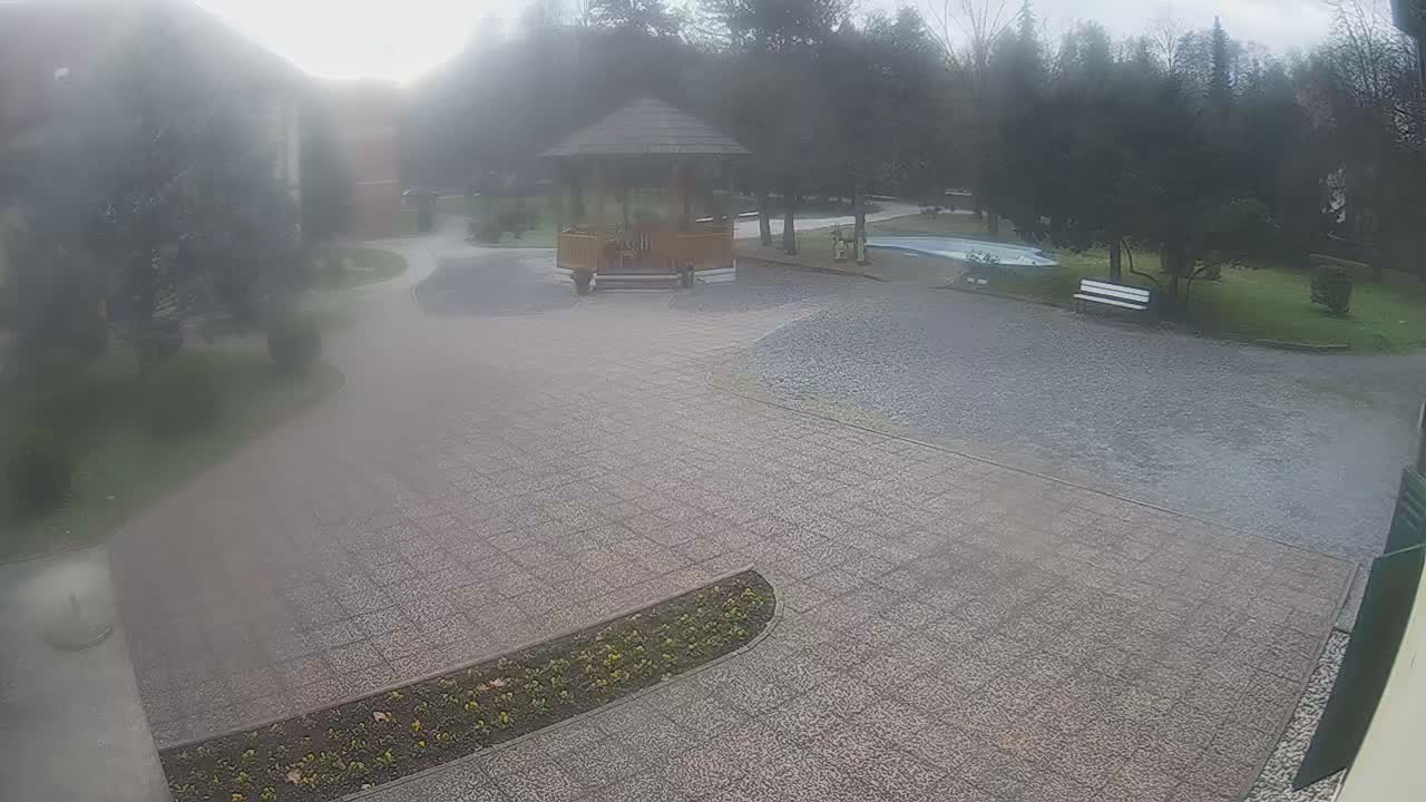 An outdoor scene on an overcast and hazy day shows a paved courtyard leading to a wooden gazebo, surrounded by trees, green grass, a white bench, and a small pool.