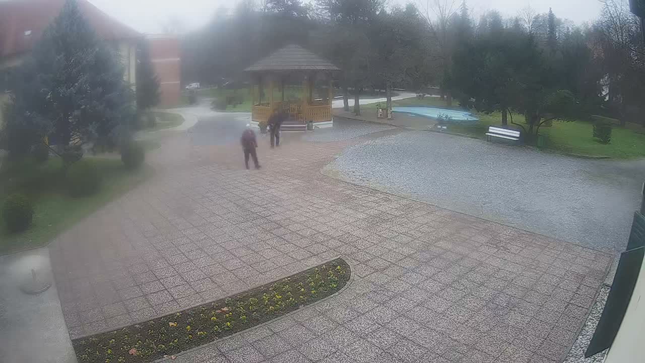 On an overcast and rainy day, two figures walk across a wet paved courtyard towards a wooden gazebo, surrounded by trees, a light blue pond, and distant buildings.
