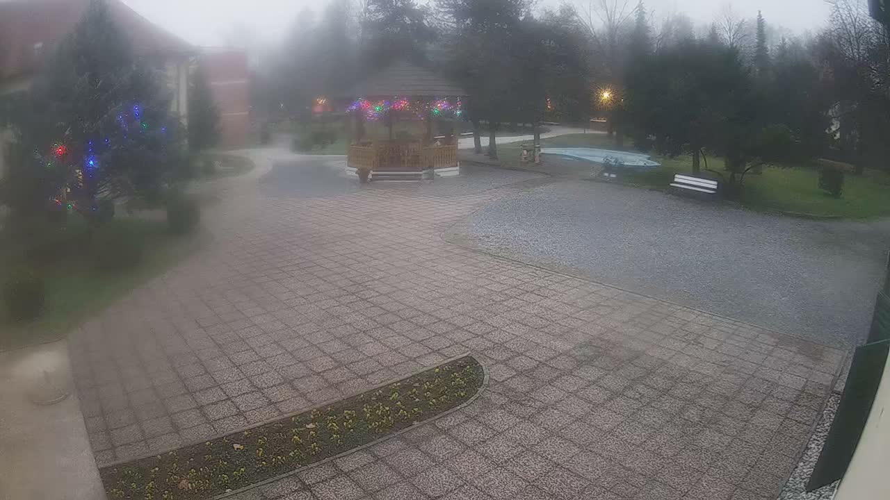 On an overcast and rainy day, two figures walk across a wet paved courtyard towards a wooden gazebo, surrounded by trees, a light blue pond, and distant buildings.