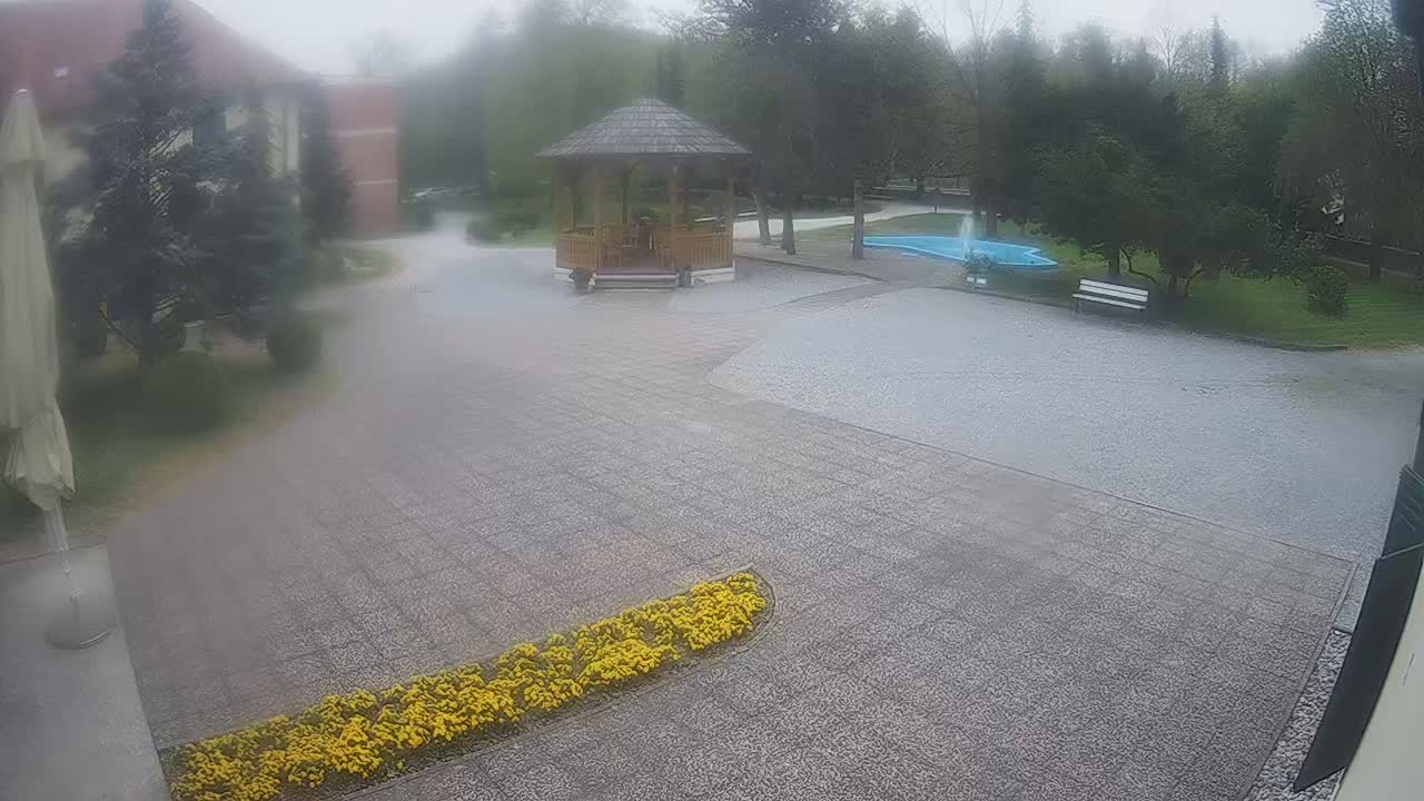 On an overcast and rainy day, two figures walk across a wet paved courtyard towards a wooden gazebo, surrounded by trees, a light blue pond, and distant buildings.