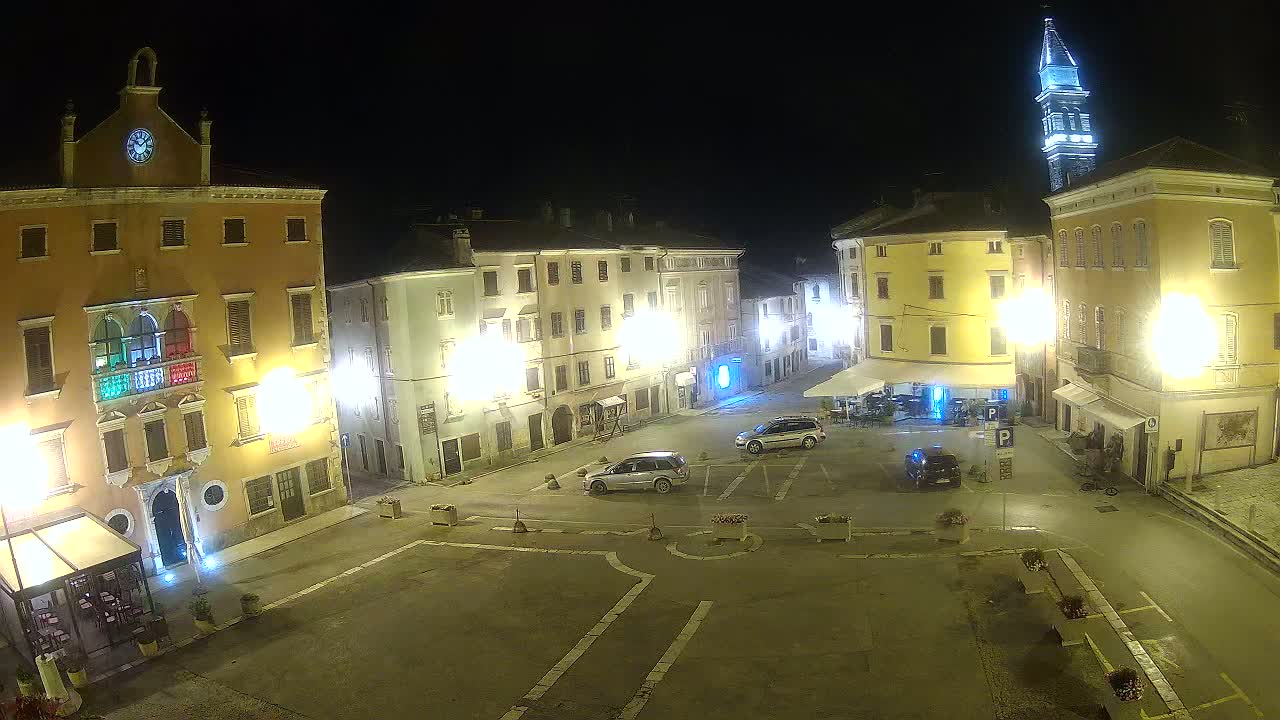 A town square at night, illuminated by streetlights, features several buildings, including a tall tower, and a few parked cars.
