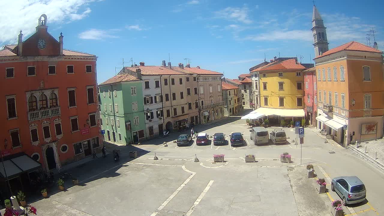 A sunny day reveals a town square with several colorful buildings, parked cars, and a few pedestrians.