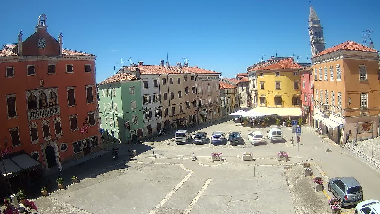 A town square with colorful buildings, parked cars, and a church steeple under a clear blue sky.