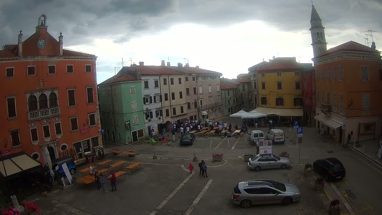 A town square with colorful buildings under a cloudy sky, filled with people gathered around tables and parked cars.
