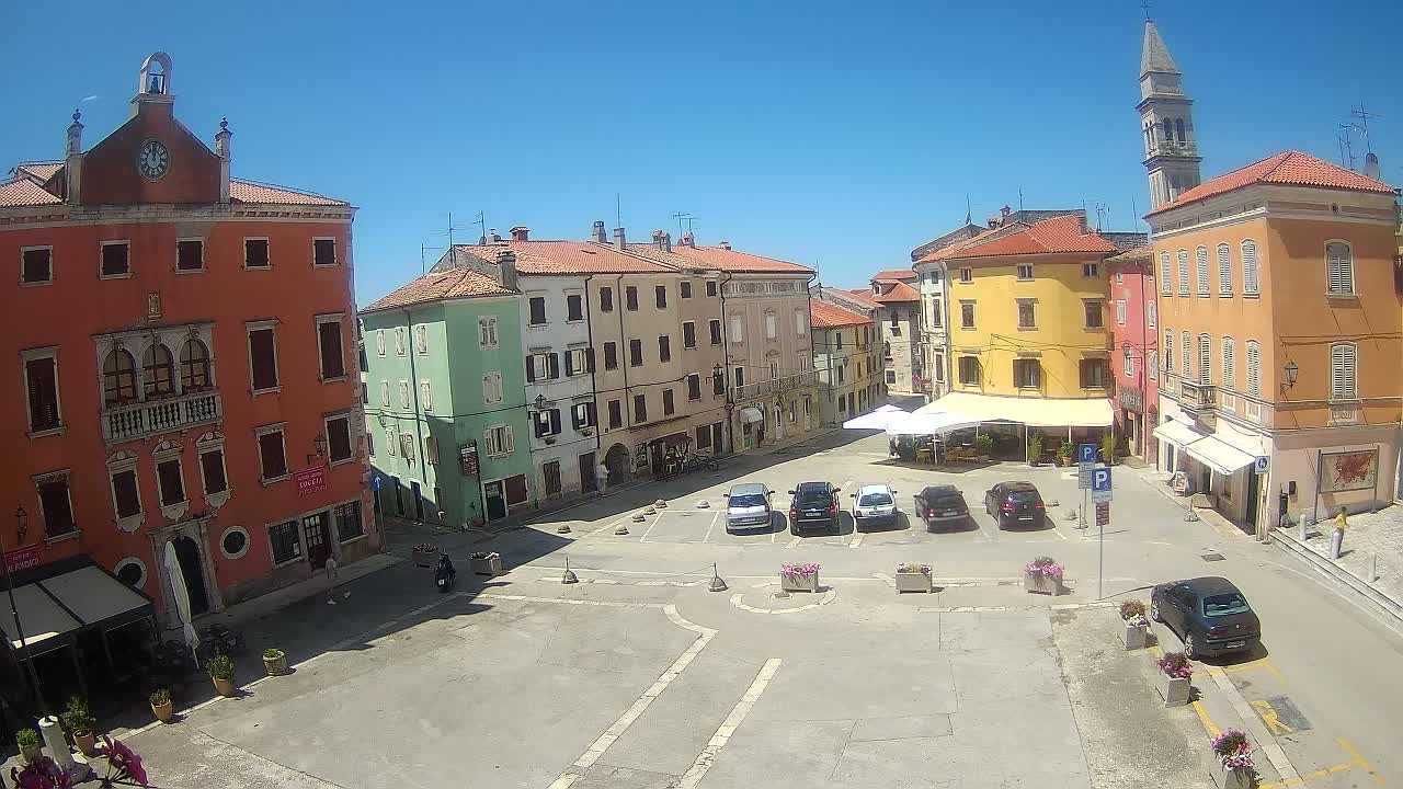 A sunny town square with colorful buildings, a few parked cars, and a tall church tower in the background.