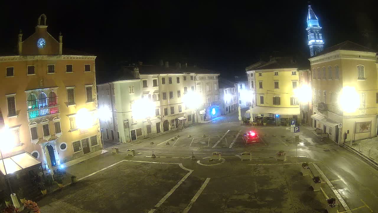 A nighttime view of a town square with several illuminated buildings, a lone vehicle, and a tall tower in the background under slightly wet conditions.
