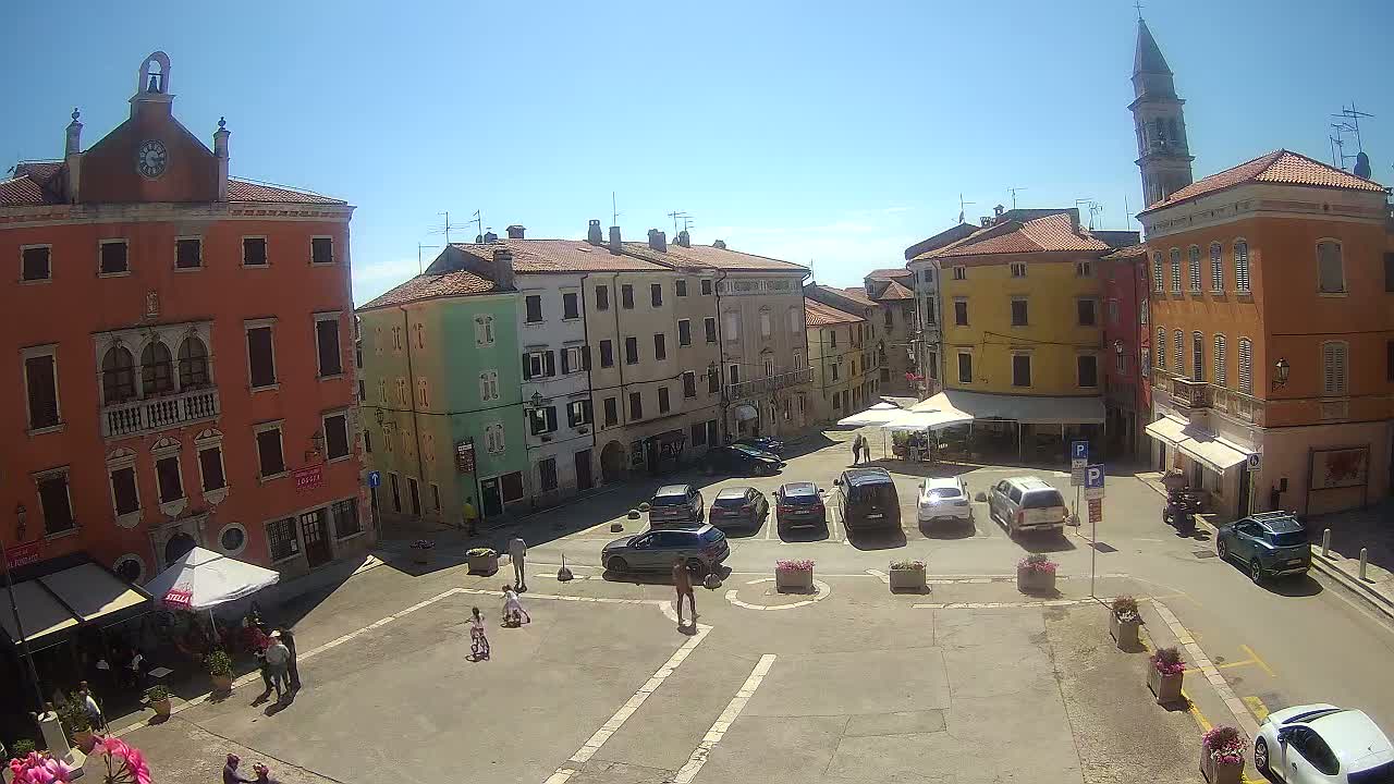 A sunny town square, surrounded by colorful buildings, with cars parked and a few people walking.