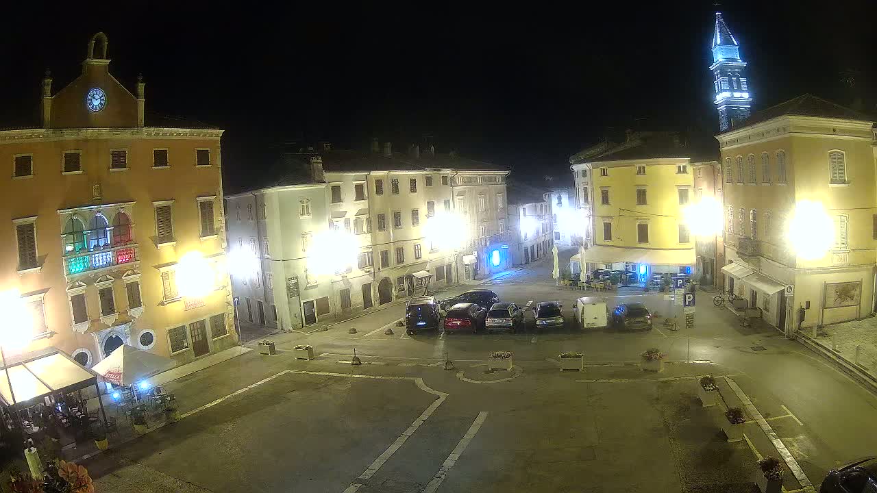 A nighttime view of a town square with several parked cars, illuminated buildings, and a tall tower in the background.