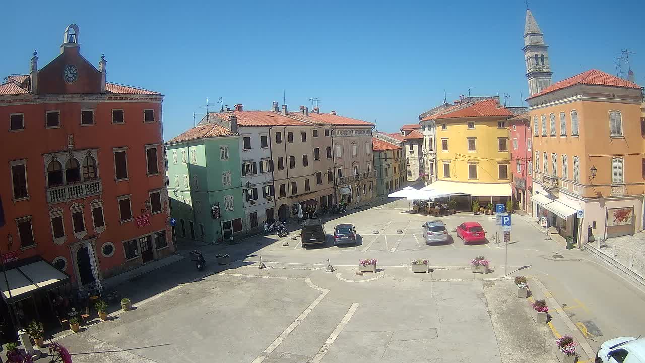 A town square with colorful buildings, a few parked cars, and a clear, sunny sky.