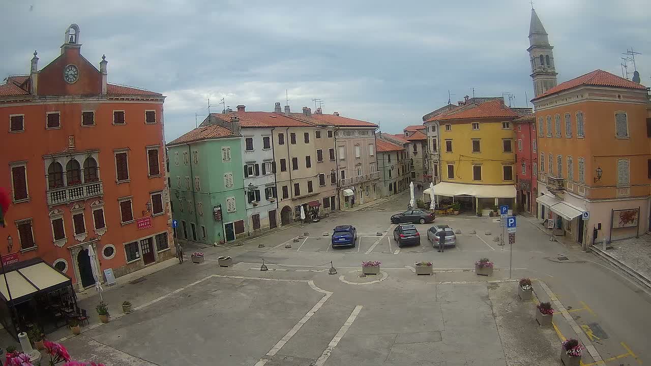 A town square with several colorful buildings surrounding a mostly empty parking area under a cloudy sky.