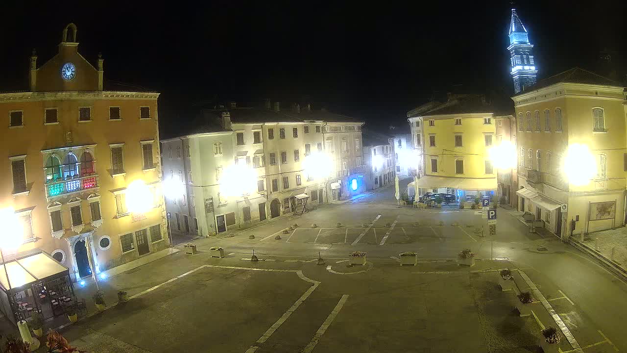 A nighttime view of a town square with several illuminated buildings surrounding a mostly empty paved area, with a tall tower visible in the background.
