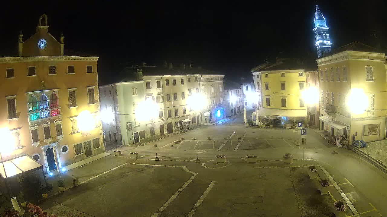 A nighttime view of an empty town square, surrounded by illuminated buildings, one of which has a clock tower.