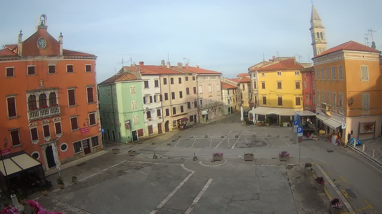 A mostly empty town square is surrounded by colorful multi-story buildings under a clear, bright sky.