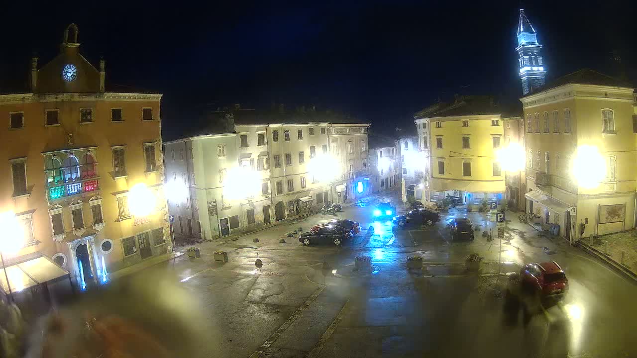 A wet town square at night, illuminated by streetlights, features several parked cars and buildings with tall windows, one of which has a clock tower.