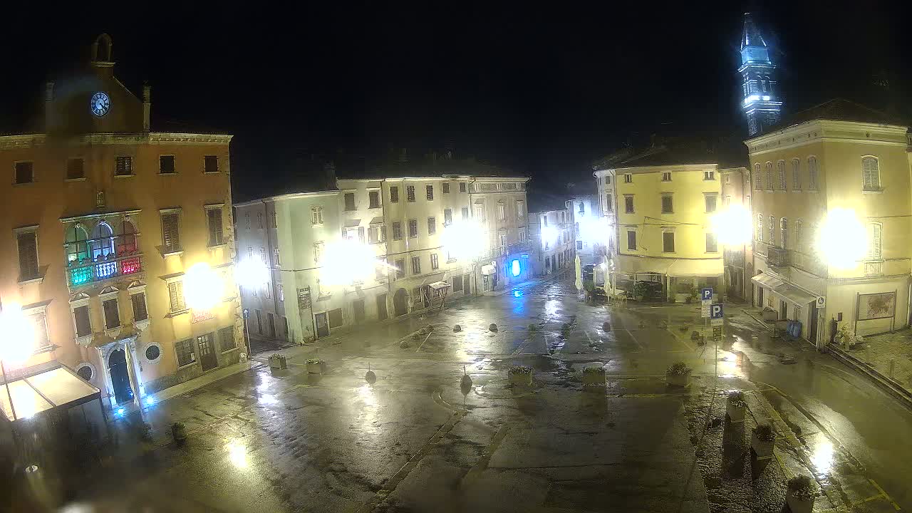 A wet, illuminated town square at night, surrounded by buildings, one with a tall tower.