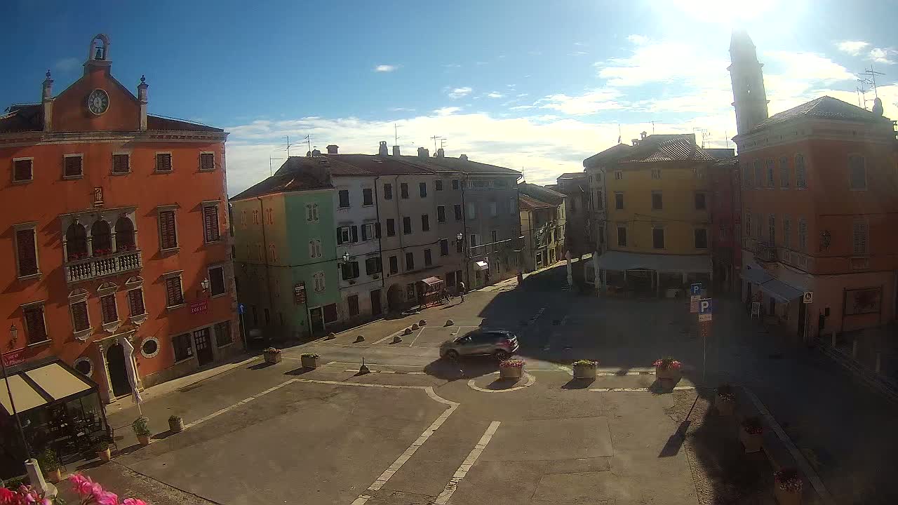 A sunny day reveals a town square surrounded by multi-storied buildings of varying colors, one car is visible in the square.