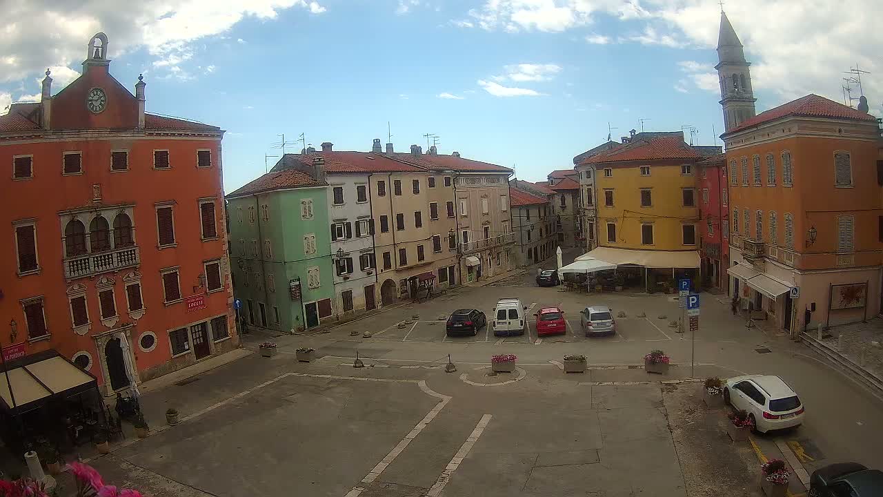 A town square with several multi-story buildings in various pastel colors, a few parked cars, and partly cloudy skies.