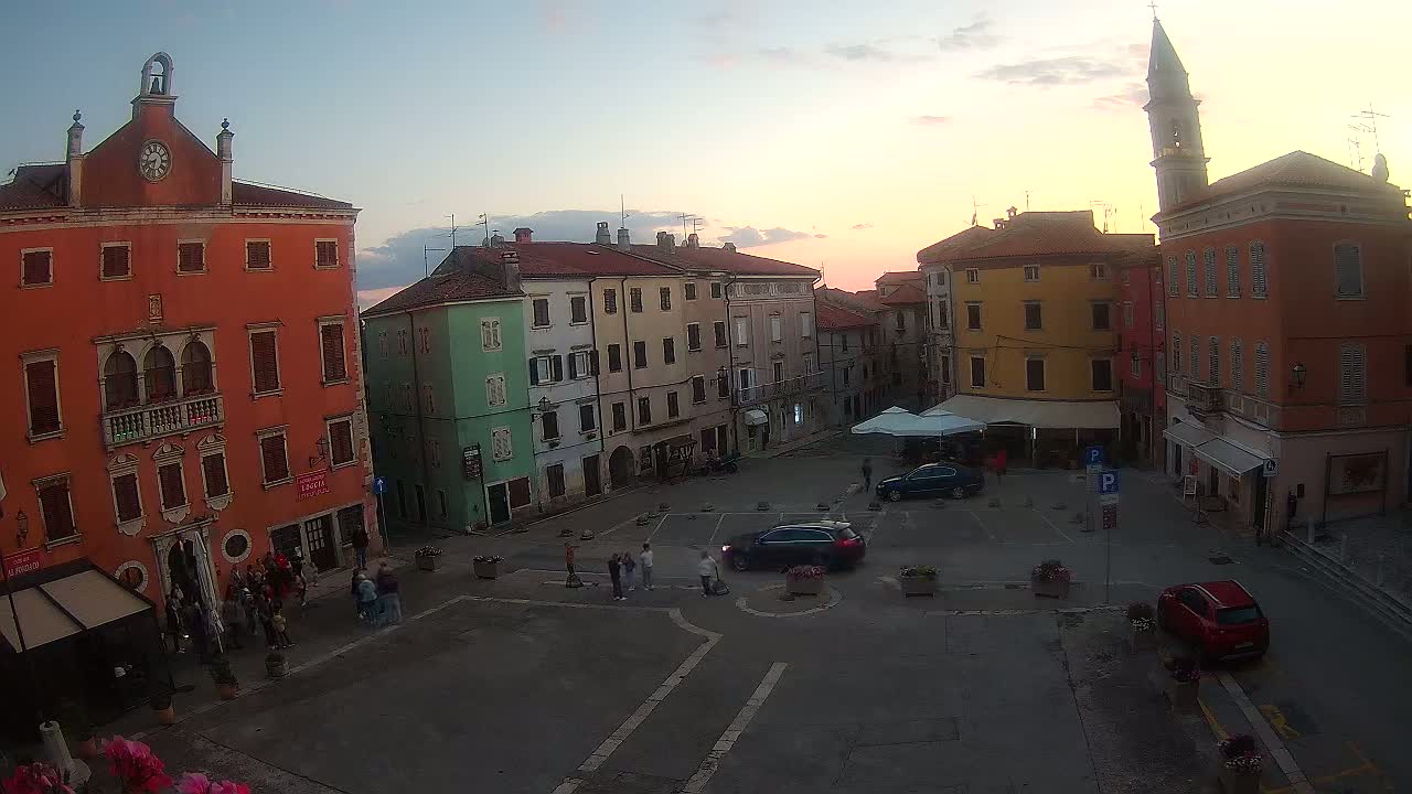 A town square with colorful buildings at sunset, a few people milling about, and several cars parked.