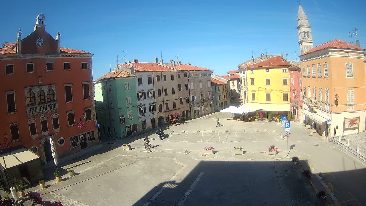 A mostly empty town square is surrounded by colorful, multi-story buildings on a sunny day.