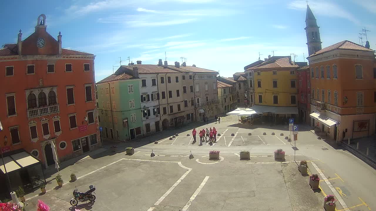 A sunny day reveals a town square surrounded by colorful buildings, a small group of people standing near the center, and a motorcycle parked near the bottom.