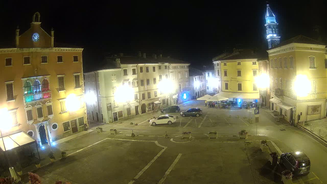 A nighttime, brightly lit town square with several multi-story buildings, a few cars parked, and a tall tower in the background.