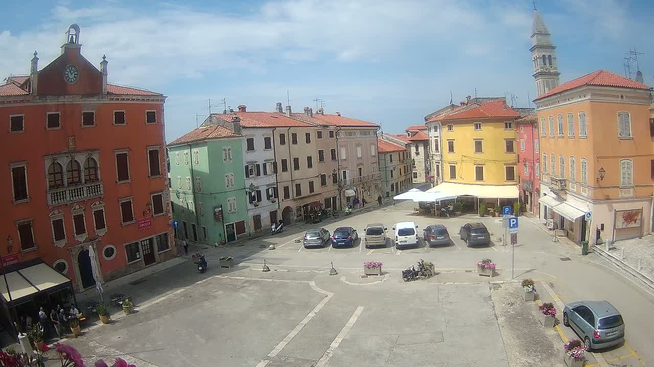 A town square with colorful buildings, parked cars, and a partly sunny sky.