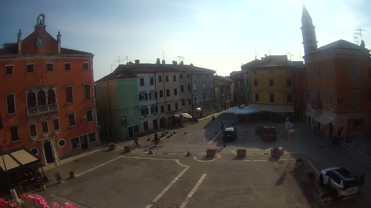 A sunny day reveals a town square with several multi-story buildings, a few cars, and some cyclists.