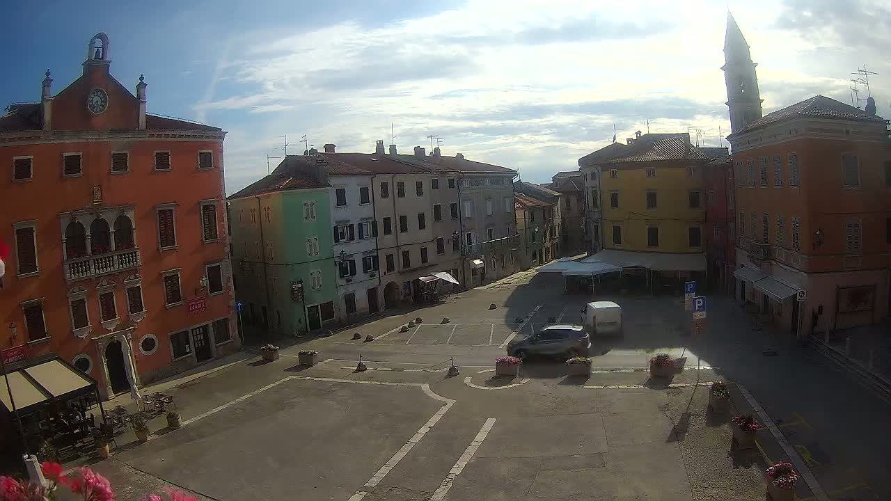 A sunny day reveals a town square surrounded by colorful buildings, including a tall orange building with a clock tower, and a few cars parked in the square.