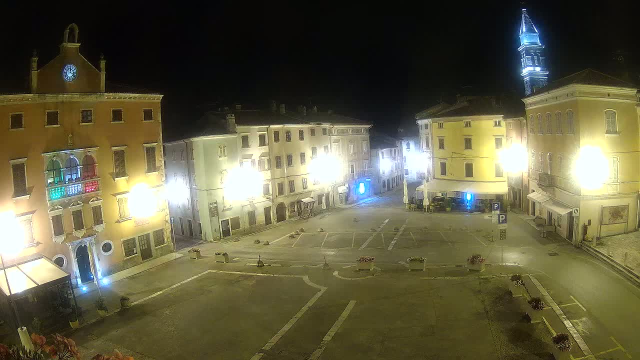 A nighttime view of a town square with several brightly lit buildings surrounding a mostly empty paved area, including a tall tower.