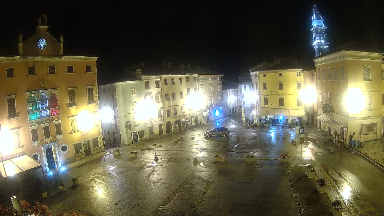 A wet, illuminated town square at night, surrounded by multi-story buildings, one of which has a clock tower.