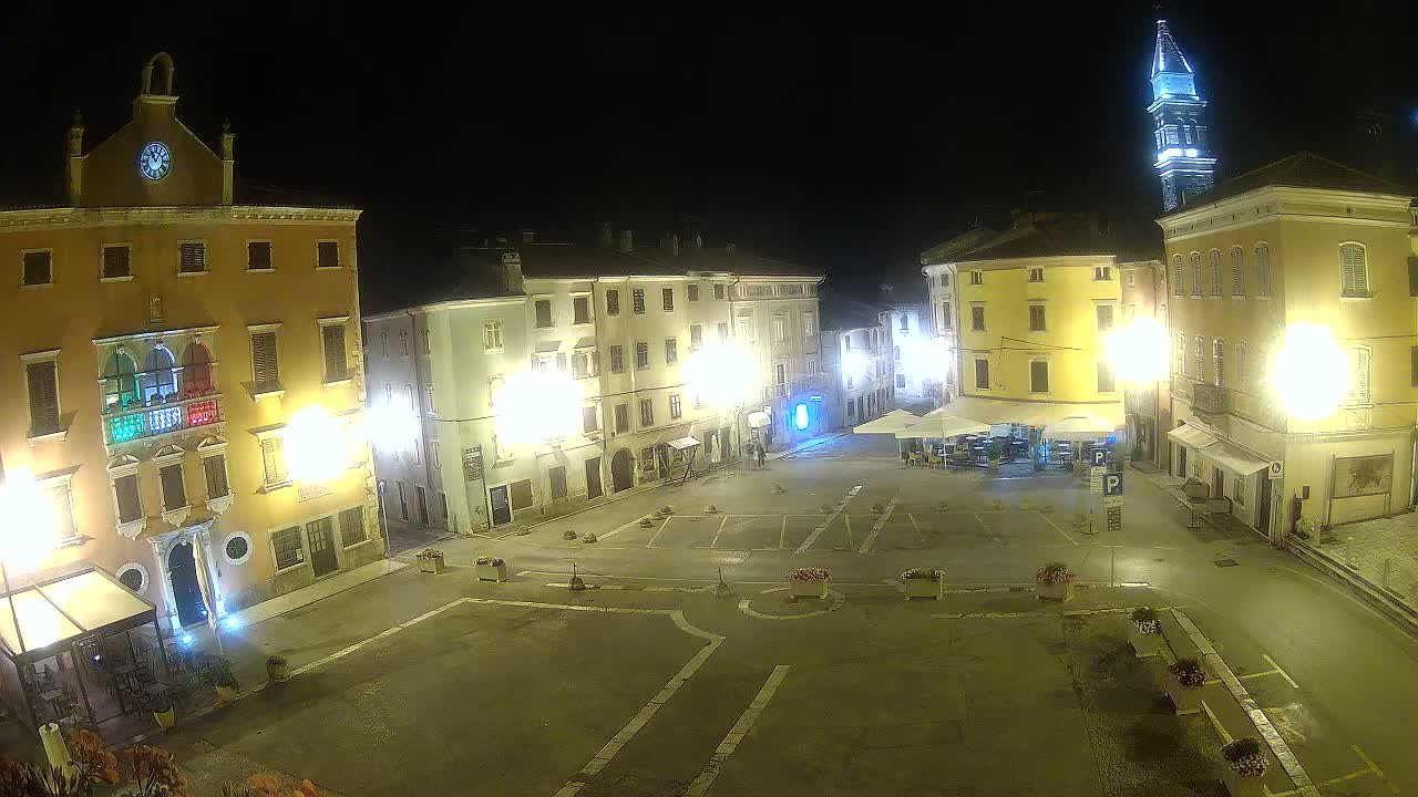 An empty town square at night, illuminated by streetlights, is surrounded by multi-story buildings, one of which has a tall tower.