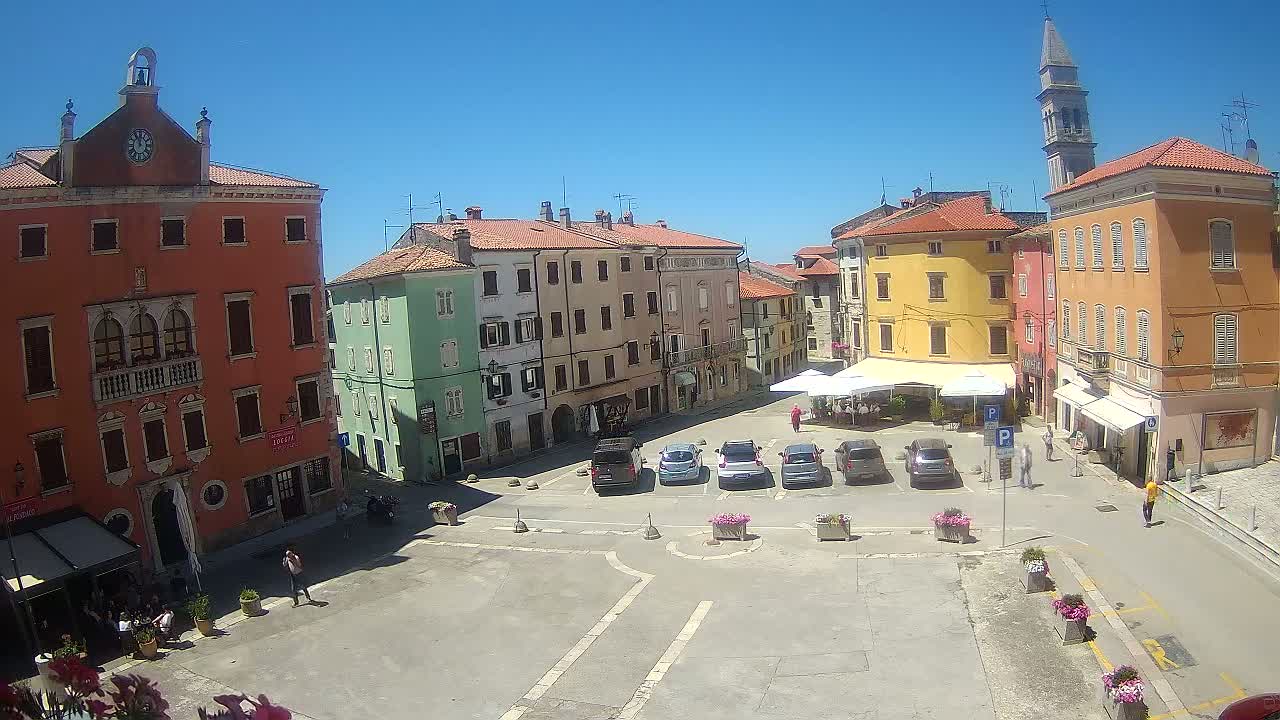 A sunny day reveals a town square with colorful buildings, parked cars, and a few pedestrians, all under a clear blue sky.