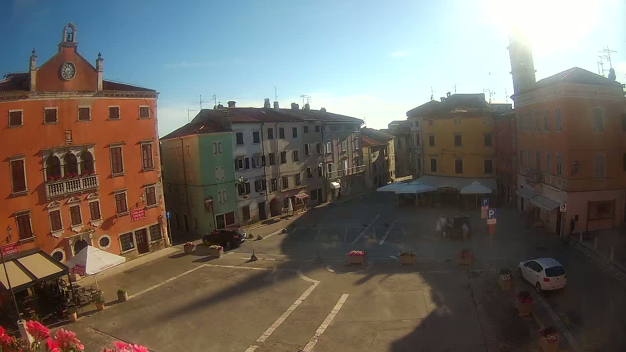 A sunny day reveals a town square surrounded by colorful buildings, some with visible balconies and clock towers, with a few cars and people visible.