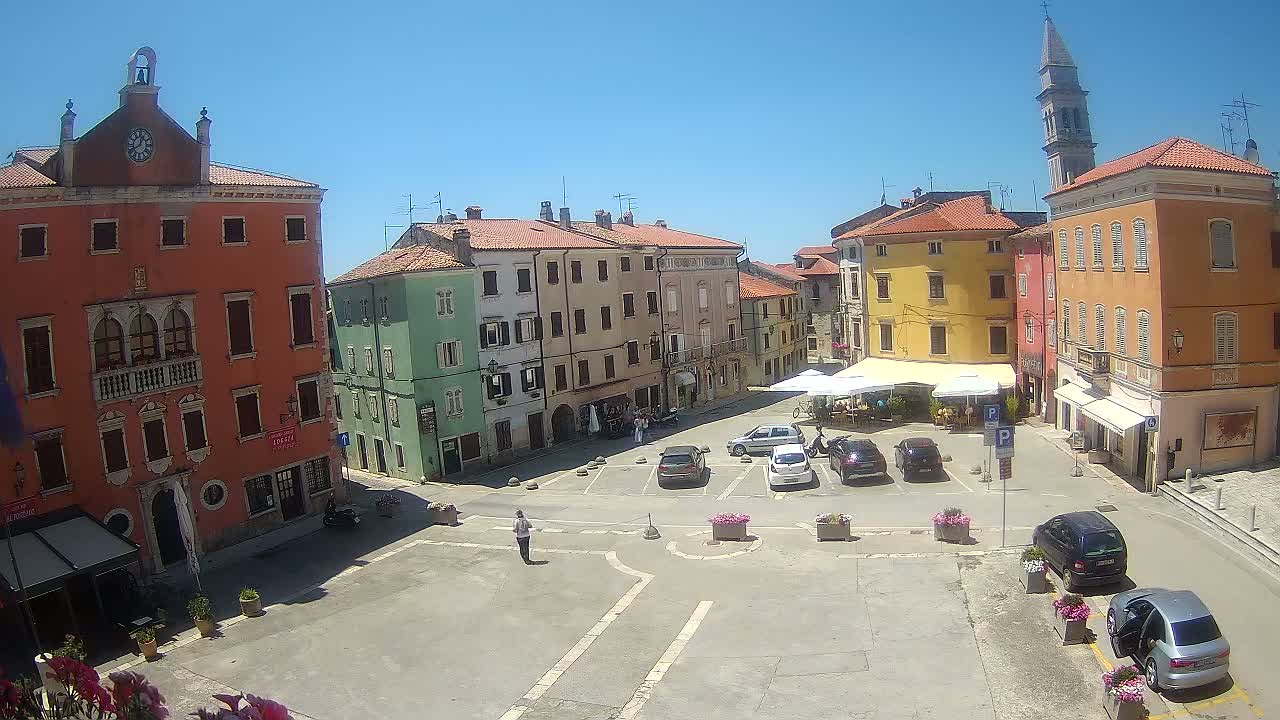 A sunny town square with colorful buildings, parked cars, and a few people walking around.