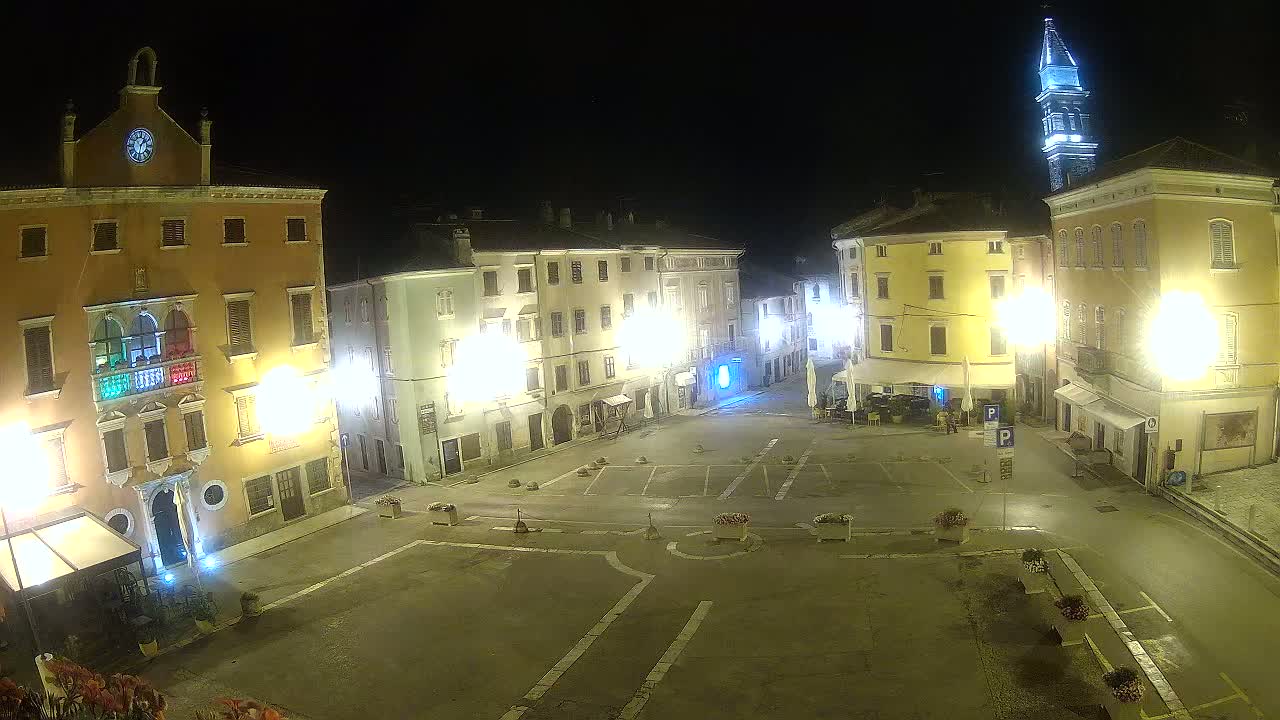 An empty town square at night, illuminated by streetlights, is surrounded by brightly lit buildings, one of which has a clock tower.