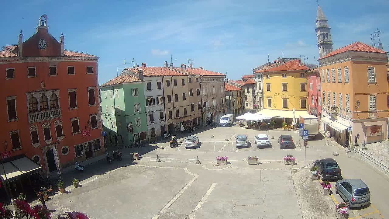 A sunny day in a town square with colorful buildings, parked cars, and a few people.