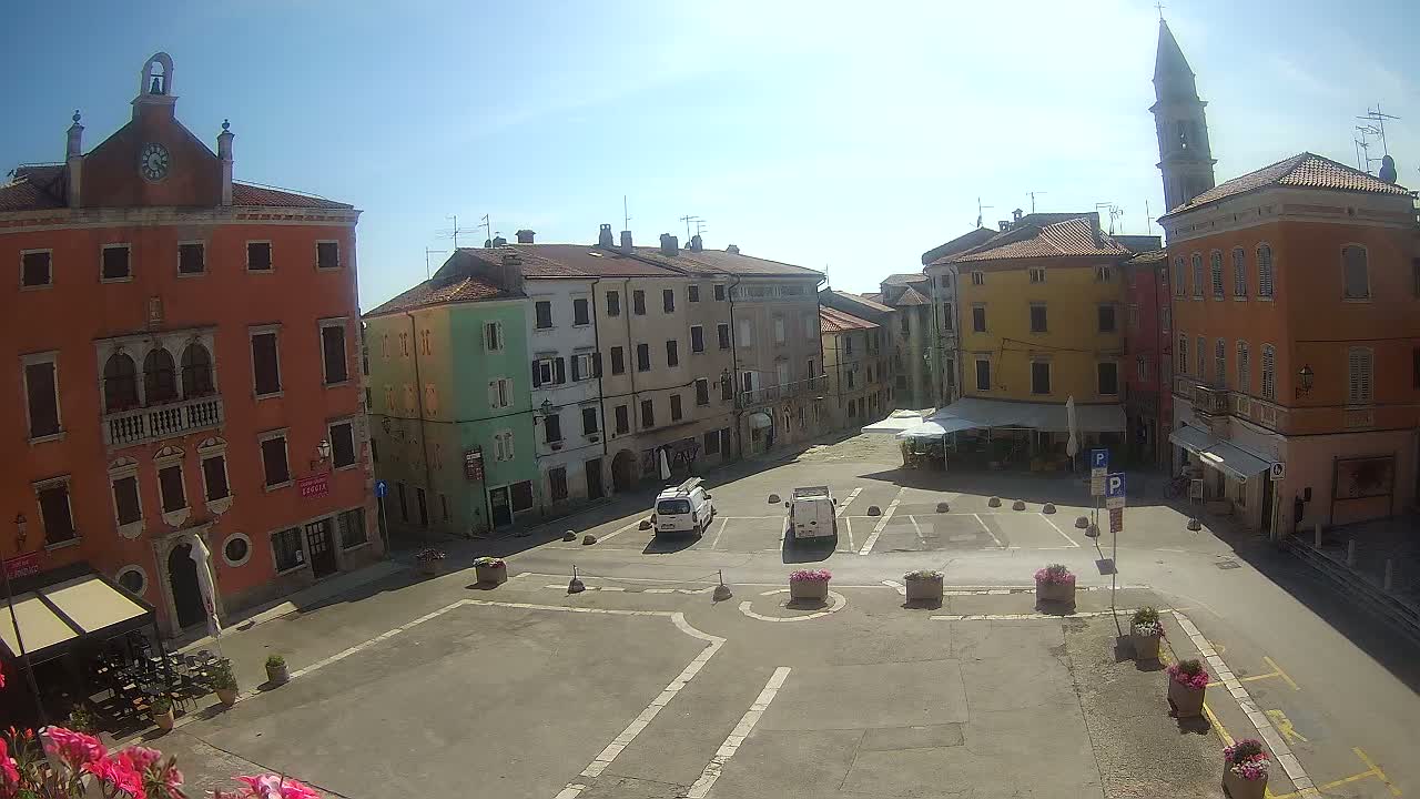 A sunny town square is seen from above, featuring several multi-story buildings in various pastel shades, with a few vehicles parked in designated spaces.