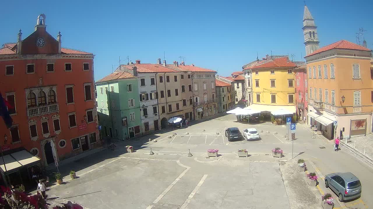 A town square with colorful buildings, a few parked cars, and a bell tower under sunny skies.