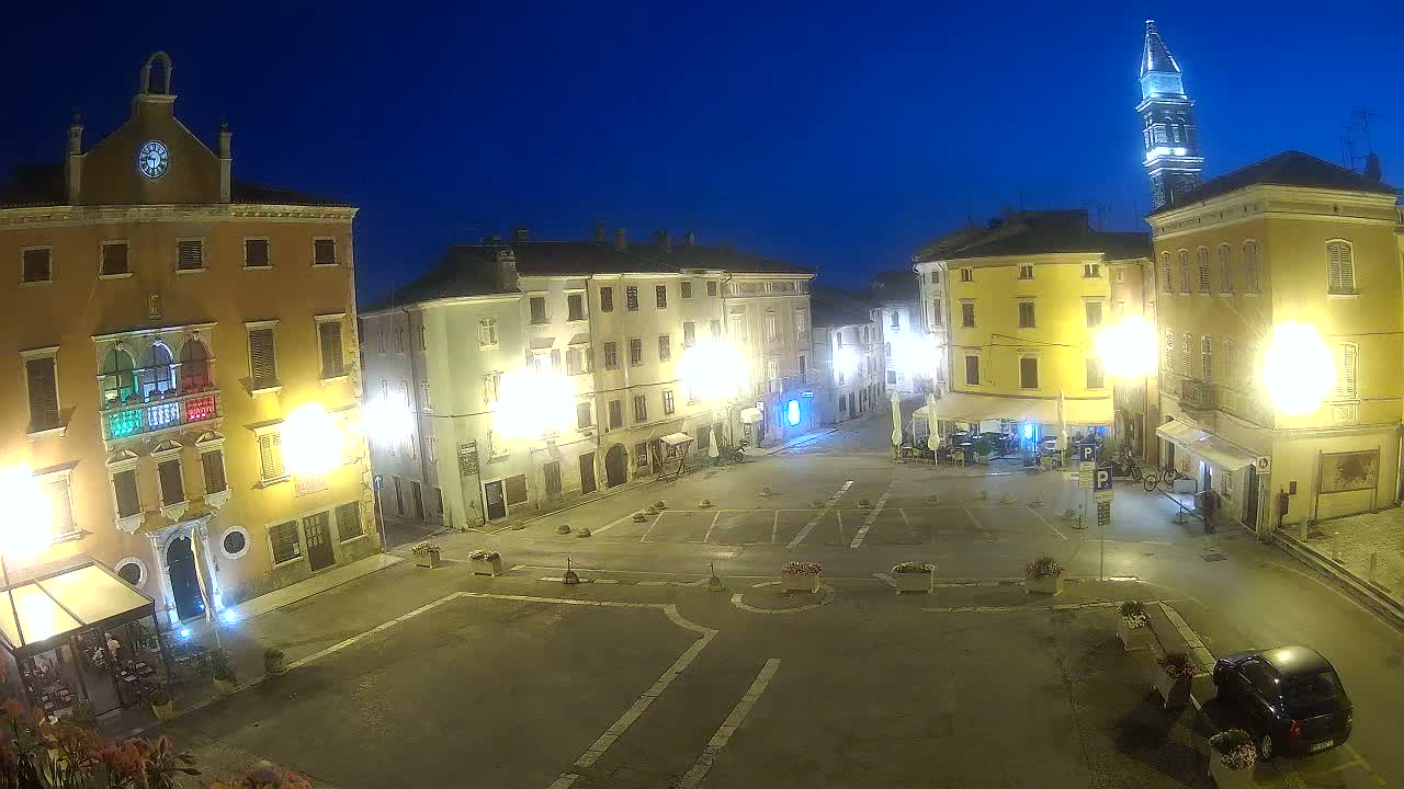 A brightly lit town square at night, under a clear, dark blue sky, is surrounded by illuminated buildings, one with a clock tower visible in the background.