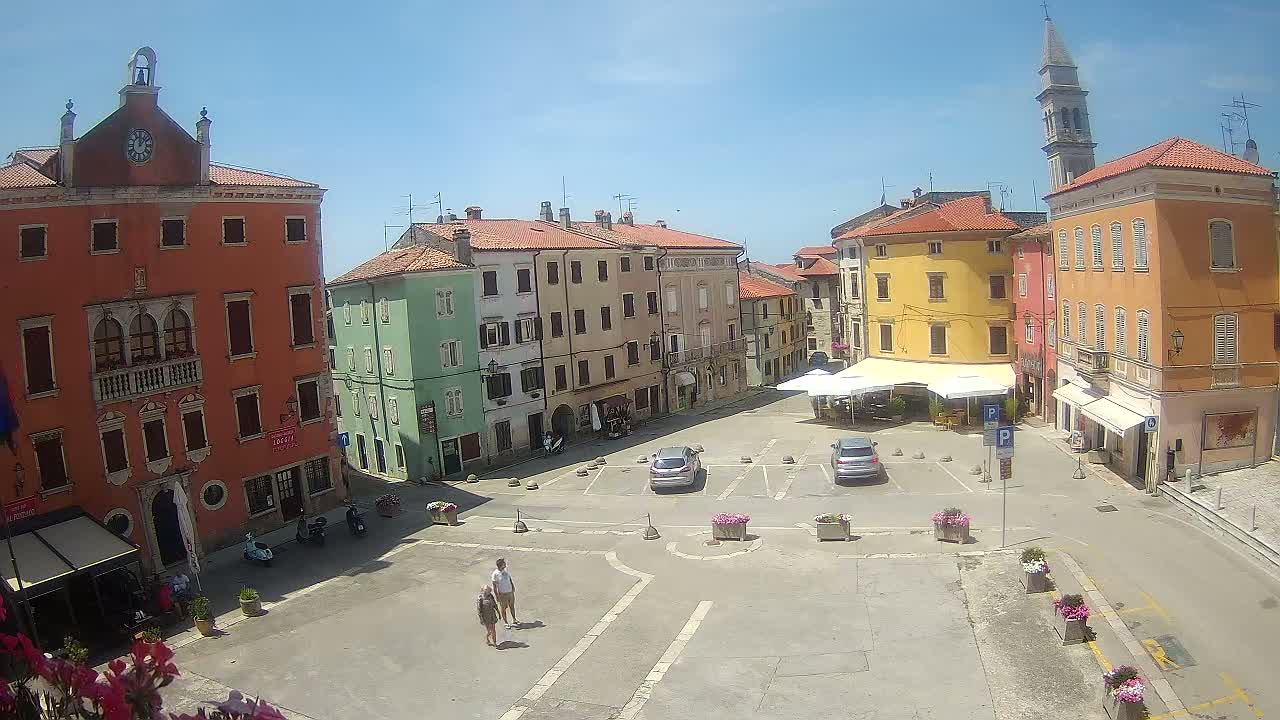 A sunny town square with colorful buildings, a few parked cars, and two people walking.
