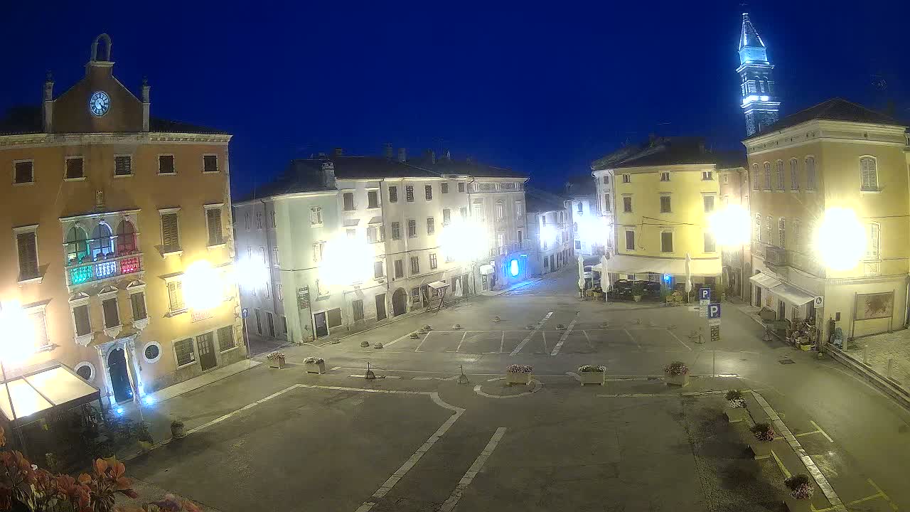 A town square at night, illuminated by streetlights, is surrounded by buildings, one of which has a tall tower.